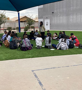 Students sitting outside on grass with adult standing in the middle of them